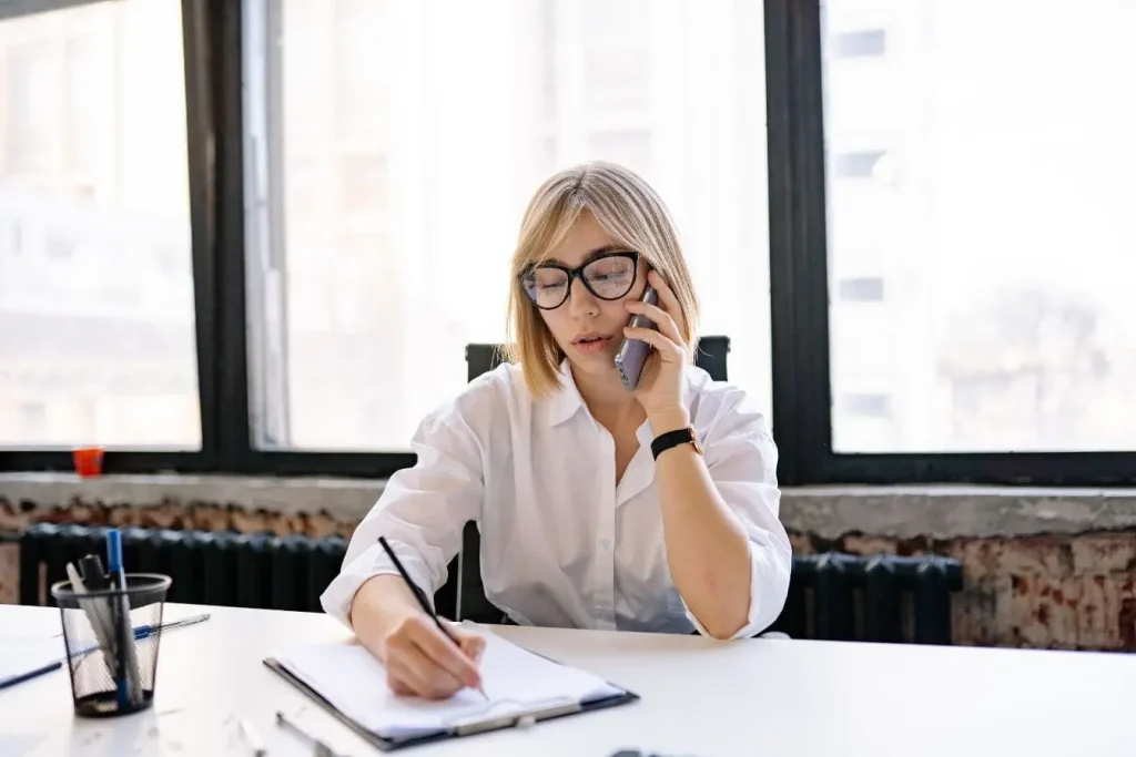 Personne en chemise blanche au bureau parlant au téléphone tout en écrivant, image qui illustre la préparation d’une proposition commerciale.