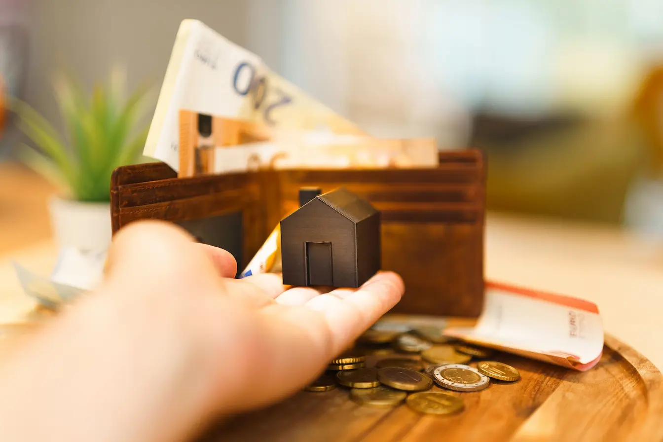 Main tenant une petite maison noire devant portefeuille rempli de billets, symbole du financement participatif en immobilier.