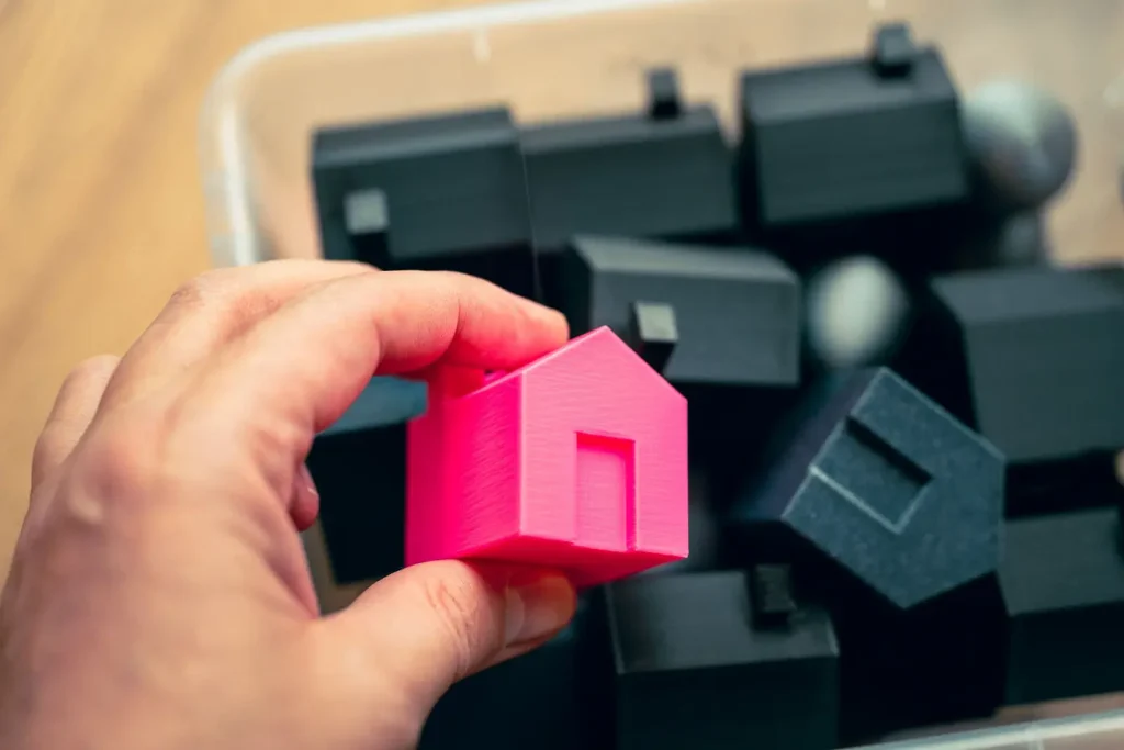 Main tenant une maison rose miniature devant plusieurs modèles noirs, symbole du choix unique offert par le crowdfunding immobilier.