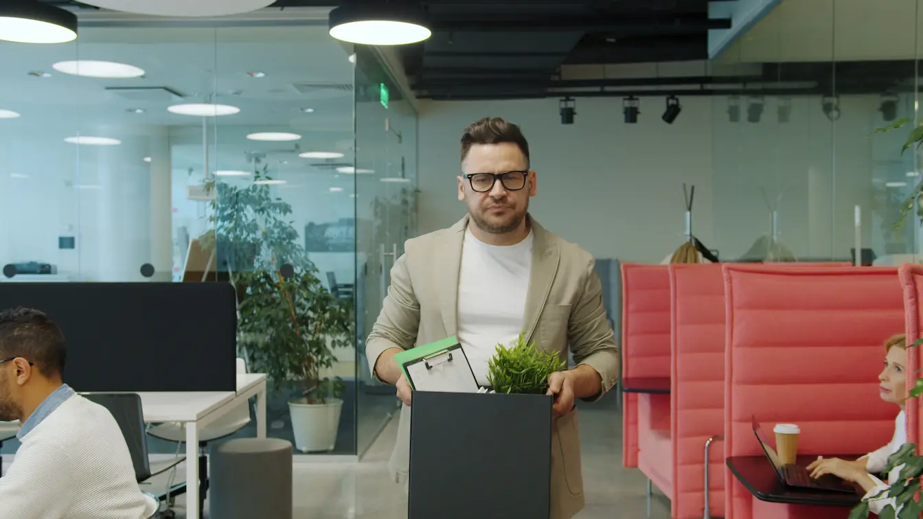 Employé en blazer beige Employé en blazer beige portant carton avec plante et dossiers, symbole des départs professionnels liés à rupture conventionnelle ou démission.